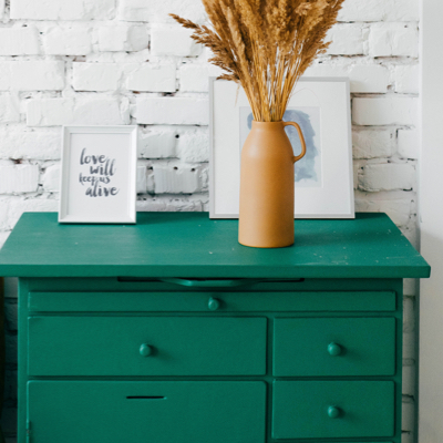 Green dresser against white brick wall with two picture frames and a decorative vase.
